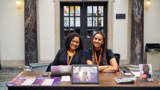 Two black women sat behind a desk looking at the camera